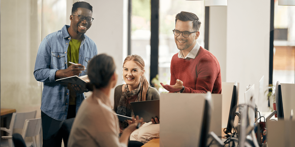 A group of smiling people sit and stand around a colleague's desk as they brainstorm ideas for building a learning culture in their accounting firm.