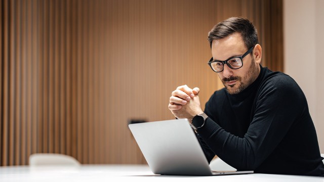 A man in glasses, wearing a black shirt, looks pensively at a laptop with his hands folded together in front.