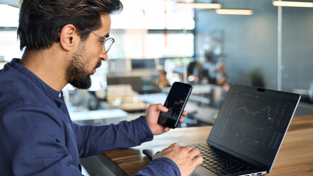 A man wearing glasses and a long-sleeved navy shirt, looks at both a laptop and his smartphone as he monitors the blockchain.