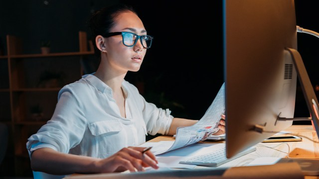 A woman wearing glasses looks at a monitor with a newspaper in one hand.