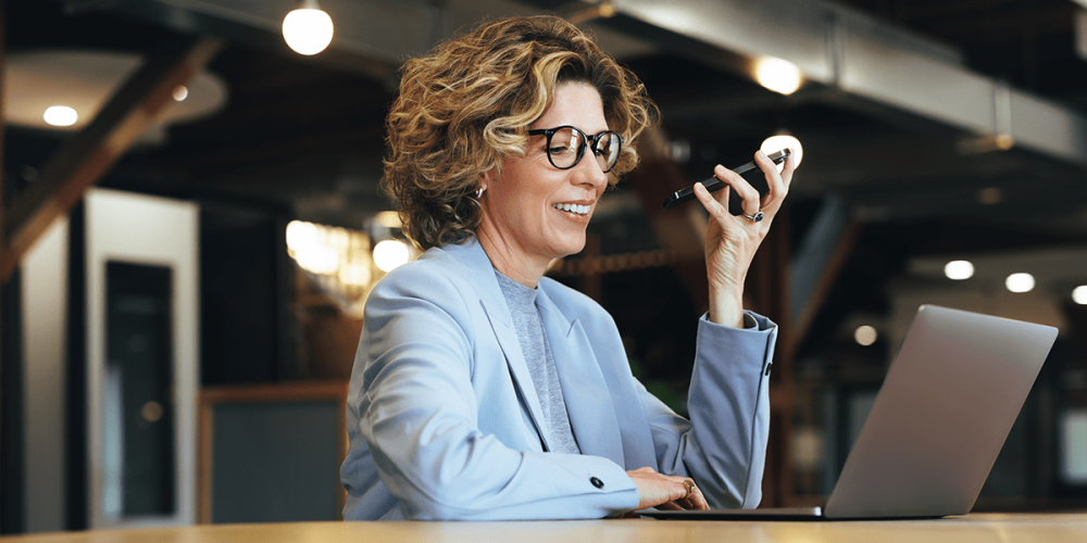 A woman with glasses holds her smartphone in her left hand while looking at her laptop.