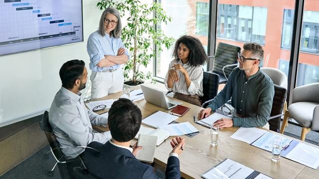 Accounting firm clients sit around a conference room table, listening to a firm leader.