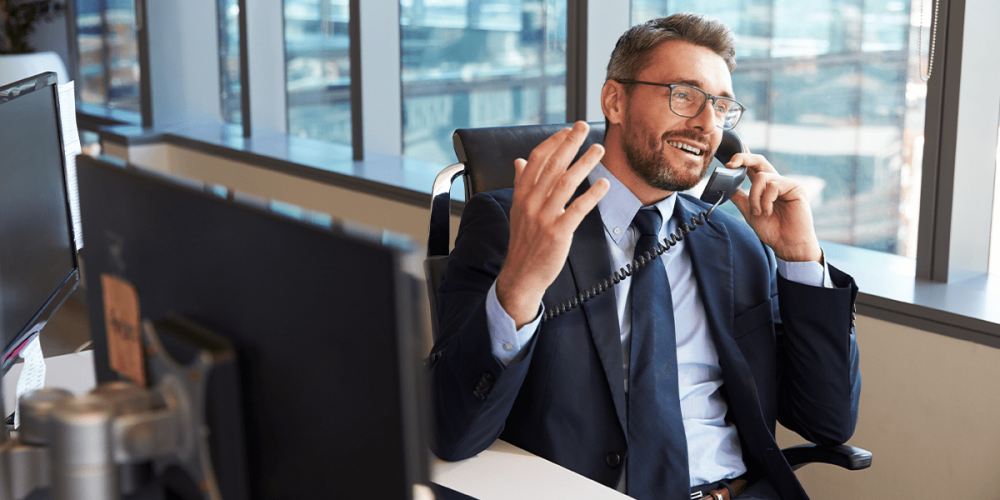 A male accountant sits at a desk in an office, talking on the phone about achieving accounting firm growth through recurring revenue strategies.