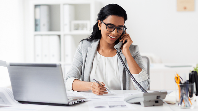 A woman sits at a desk in front of a laptop, talking on the phone to a client about advisory services.