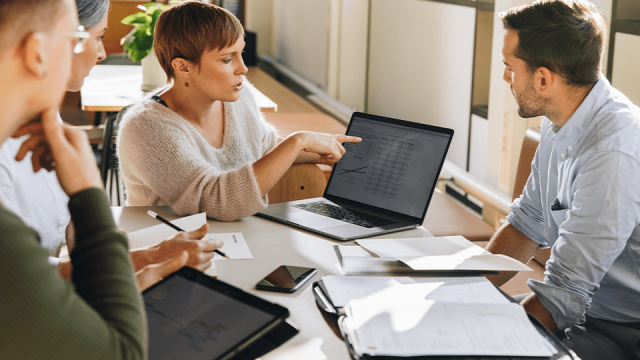 A woman points to a laptop screen while male coworkers look on.