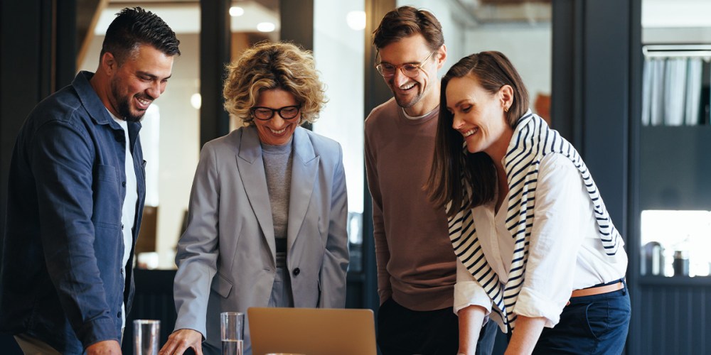 A group of four people stand smiling around a laptop as they discuss more effective communication and collaboration strategies.