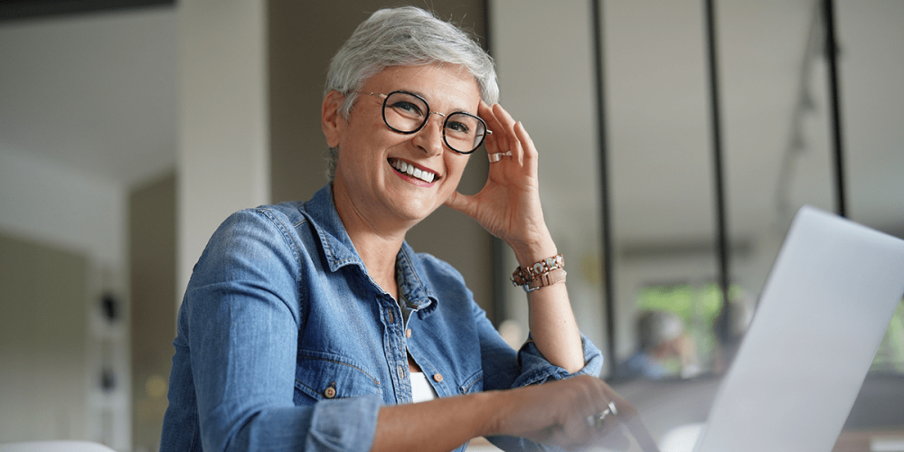 A smiling woman sits at a laptop as she contributes to an asynchronous meeting that fits her schedule.