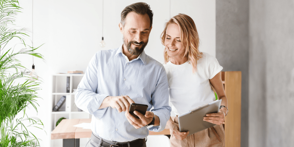 A man and woman stand smiling while they look at a smartphone.