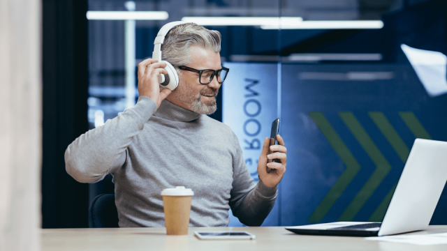 A man wearing headphones sits at a desk, looking at a laptop and holding his smartphone in one hand as he listens to an accounting podcast.