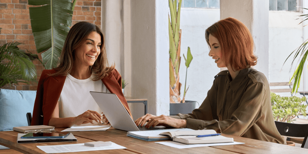 Two smiling women sit at a conference table in an office, going over the new employee onboarding plan.