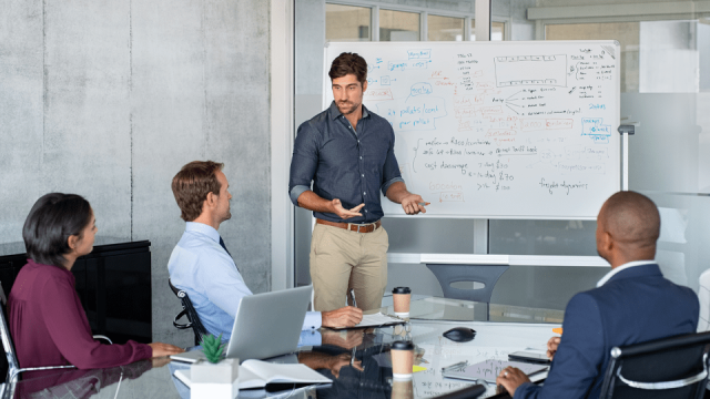 A man stands in front of a whiteboard, educating his colleagues.