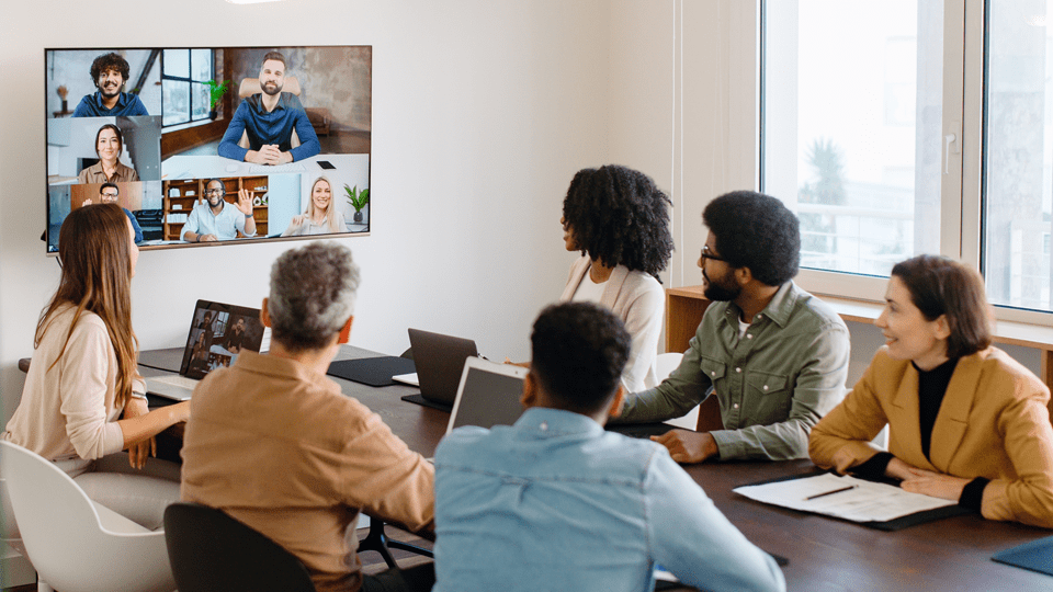 An image of a hybrid work environment with team members gathered around a conference table, interacting with other team members on a video conference.