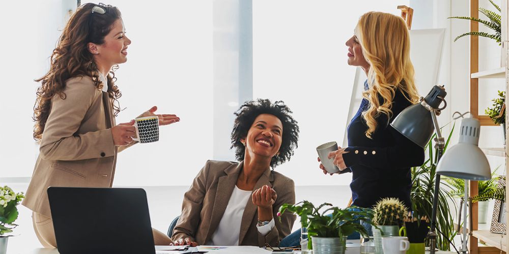 A woman sits at a desk in an office with two women standing beside her, carrying on a conversation about how their accounting firm aligns its mission, vision and values.