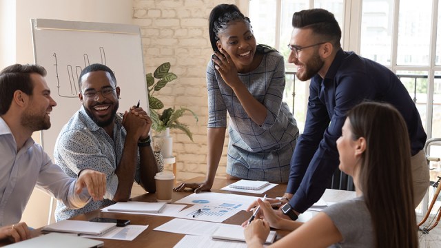 A group of people sit and stand around a conference room table, smiling as they discuss their firm's mission, vision and values.