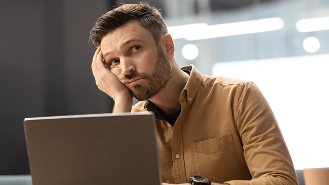 An unhappy employee sits at his desk in front of a laptop disengaged, holding his head in his hand.