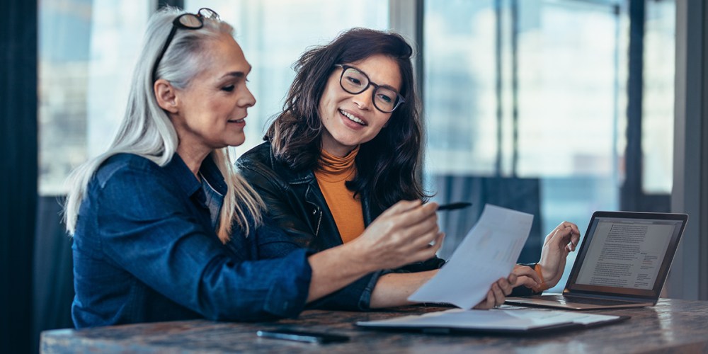 Two women sit at a conference table with a laptop open. They both have glasses and are smiling, and one is pointing at a paper she is holding in one hand with a pen in the other as they go through an onboarding process