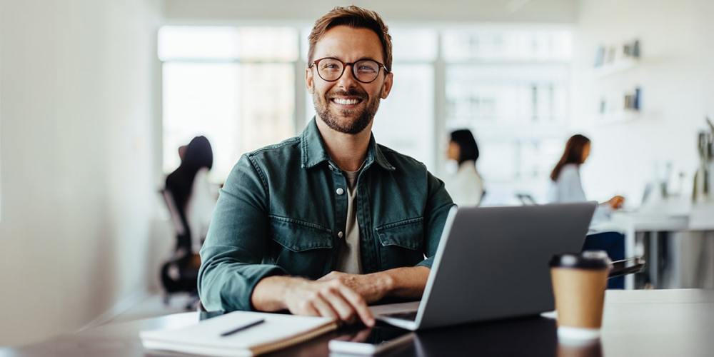 A bearded man with glasses smiles as he sits in front of a laptop because his company recognizes the link between cybersecurity and employee retention