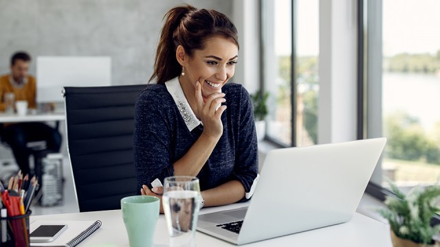 A woman sits in front of a laptop at her desk, smiling since her system runs smoothly with enough RAM.