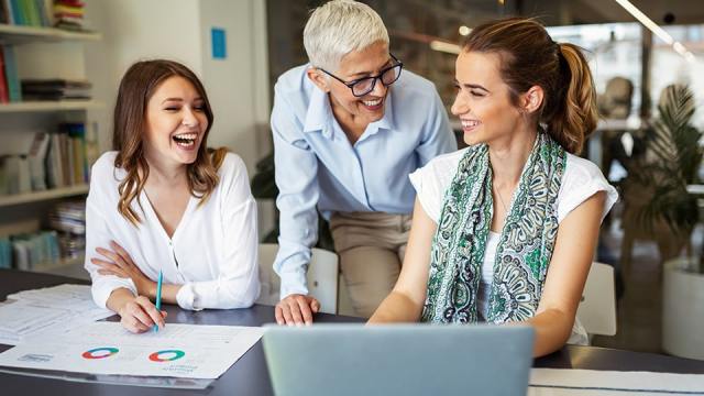 Three smiling and laughing women sit and stand at a desk because their company has created a thriving culture.