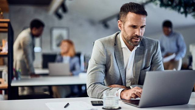 A man sits looking at tax updates from the IRS on his laptop in an office with other employees in the background during tax season.