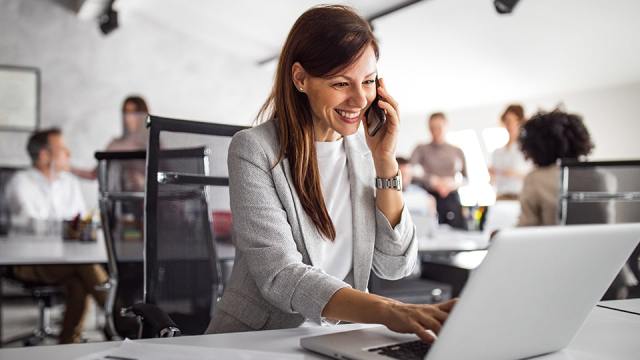 A smiling woman stands in front of a laptop as she talks on the phone.