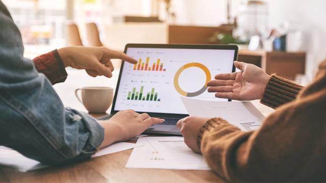 Charts and graphs appear on a laptop screen as two coworkers discuss the analytics.